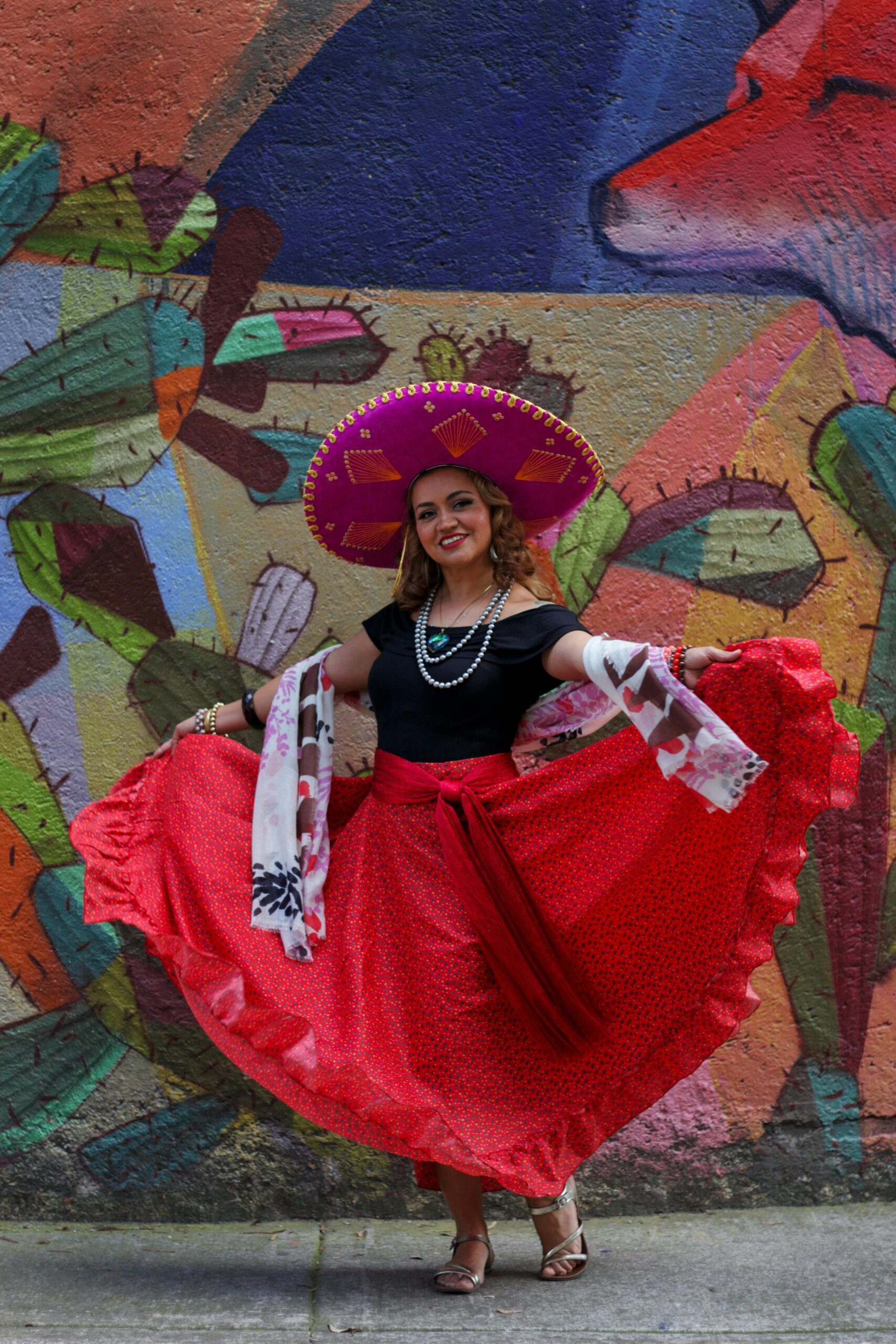 Woman in traditional Mexican dress and sombrero dancing against a colorful mural backdrop.